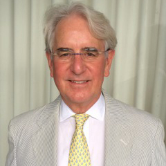 An older man with gray hair and glasses is smiling. He is wearing a light-colored suit with a white shirt and a yellow tie. The background consists of white curtains.