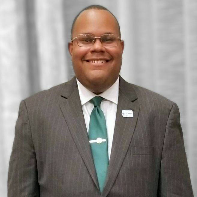 A smiling man in a suit and tie stands in front of a blurred background. He is wearing glasses and a nametag on his lapel.
