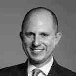 Black and white portrait of a bald man in a suit and tie, smiling slightly. The background is plain.