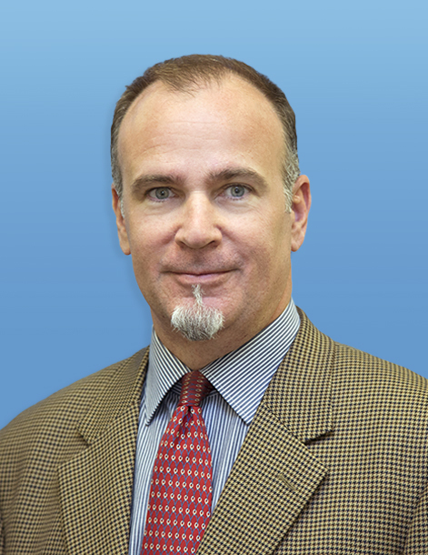 A man in a brown checked blazer, blue striped shirt, and red tie is standing against a blue background. He has short hair and a goatee.