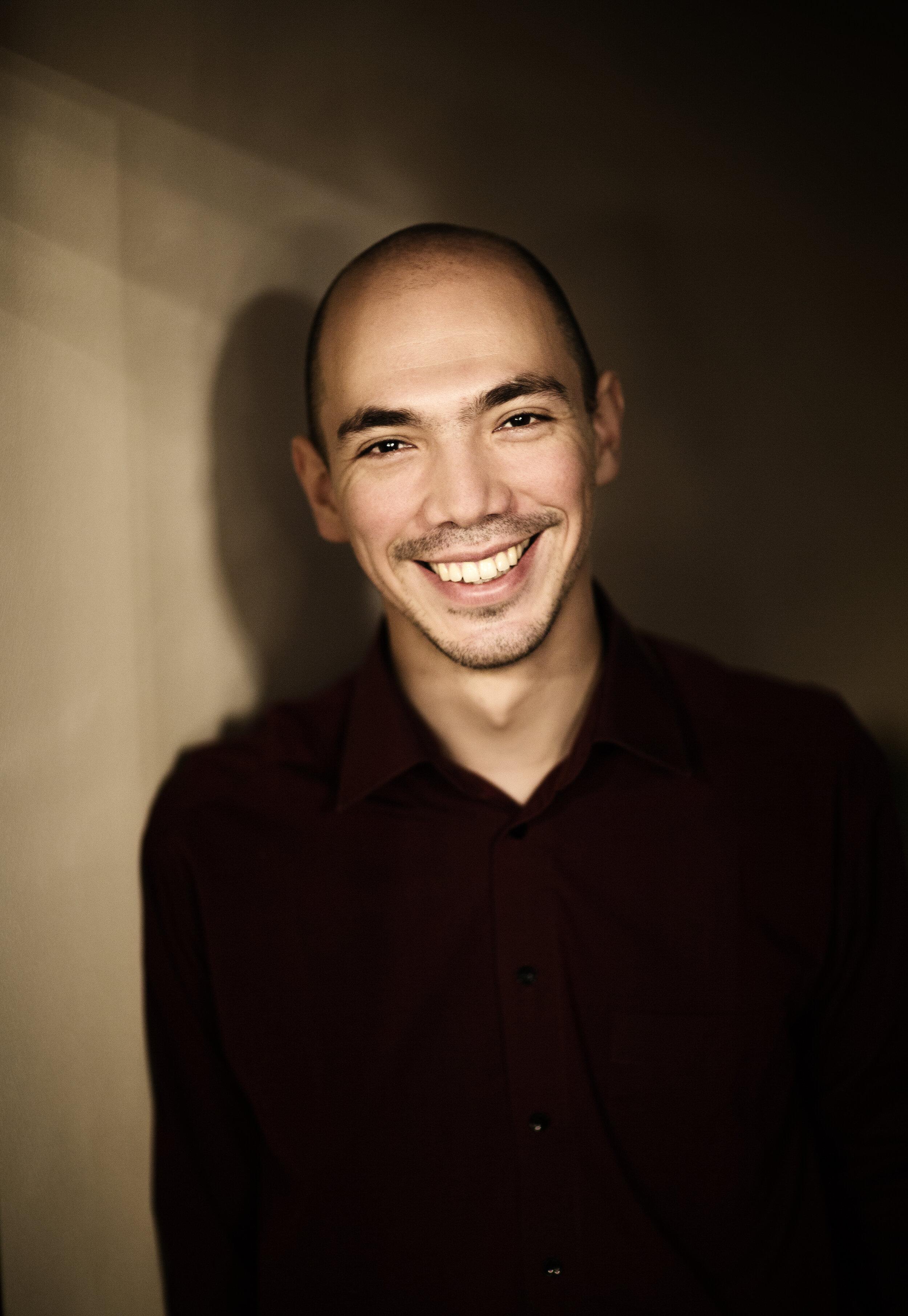 A bald man with a mustache and goatee is smiling warmly. He is wearing a dark shirt and is standing against a plain, dimly lit background.