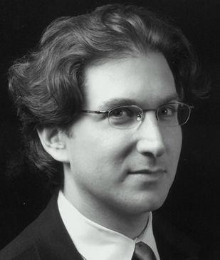 A black-and-white portrait of a person with wavy hair, wearing glasses and a suit jacket, looking slightly towards the camera. The background is dark.