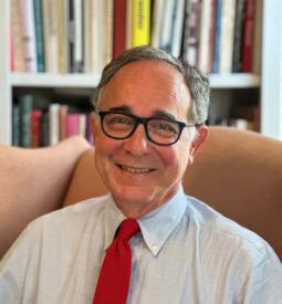 A man with glasses and gray hair is smiling while seated in an armchair. He is wearing a blue shirt and red tie. Bookshelves filled with books are visible in the background.