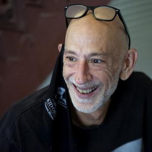 A bald man with a white beard is smiling broadly. He is wearing glasses on his head and a black shirt. The background is blurred.