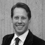 A grayscale portrait of a smiling individual wearing a suit and tie, standing against a textured wall background.