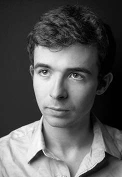 Black and white portrait of a young man with short, dark hair, wearing a light-colored shirt. He looks slightly upward and to the side, set against a dark background.