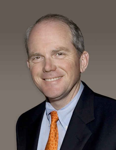 A man in a suit smiles at the camera. He is wearing a white shirt, an orange tie, and a dark jacket. The background is a neutral gray-brown gradient.