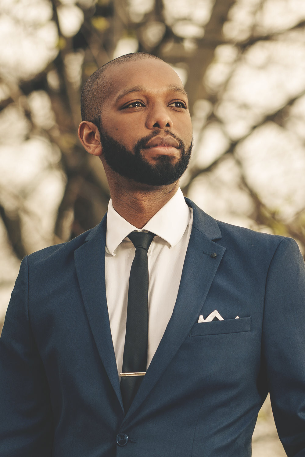 A man with a beard is wearing a blue suit, white shirt, and black tie, standing outdoors against a blurred background of bare trees. He has a thoughtful expression, and a pocket square is visible in his jacket.