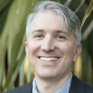 Smiling person with short gray hair stands in front of green foliage, wearing a dark jacket and light blue shirt.