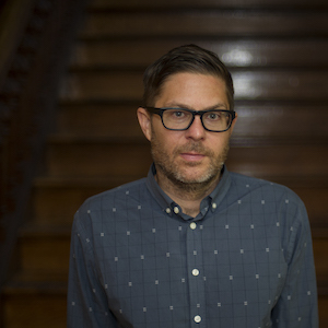 A man with glasses and a beard stands in front of a wooden staircase. He is wearing a gray, patterned button-up shirt and looking towards the camera. The background has a warm, dimly lit ambiance.