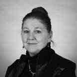 Black and white portrait of a person with their hair in a bun, wearing a collared jacket. They have a neutral expression and are looking directly at the camera. The background is plain.