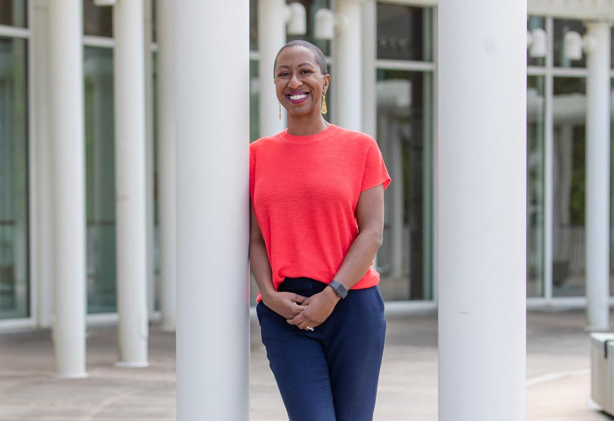 Headshot of Justene Hill Edwards in in a bright red shirt and dark pants leans against a white pillar outdoors, smiling. The background features large windows and multiple pillars.