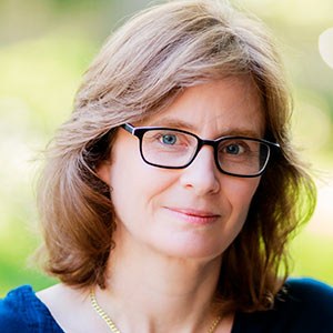 A woman with shoulder-length light brown hair, wearing glasses and a dark blue top. She is looking forward with a slight smile, set against a softly blurred green and yellow background.