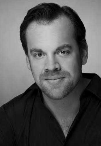 Black and white portrait of a man with short hair and a beard, wearing a dark shirt. He is looking directly at the camera with a slight smile.