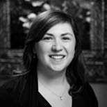 A black and white portrait of a person with shoulder-length hair, smiling at the camera. They are wearing a dark top and a necklace, with an out-of-focus, decorated background behind them.