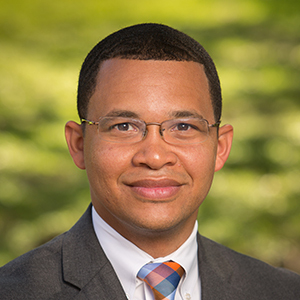 A man wearing glasses, a suit, and a multicolored tie smiles against a blurred green background.