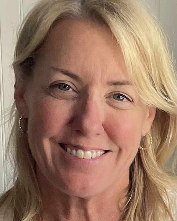 A person with light blonde hair smiles warmly at the camera, wearing hoop earrings and a light colored top. The background is softly blurred, emphasizing the subjects cheerful expression.