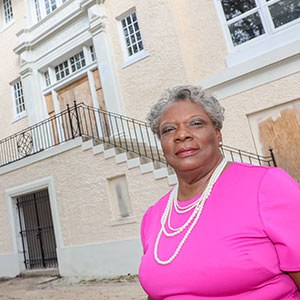 A woman in a pink dress with pearl necklaces stands outside a large beige building with white details and a staircase.