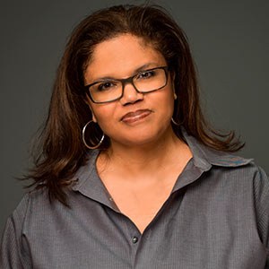 A person with long hair and glasses is wearing a button-up shirt against a dark background. They are looking directly at the camera with a neutral expression.