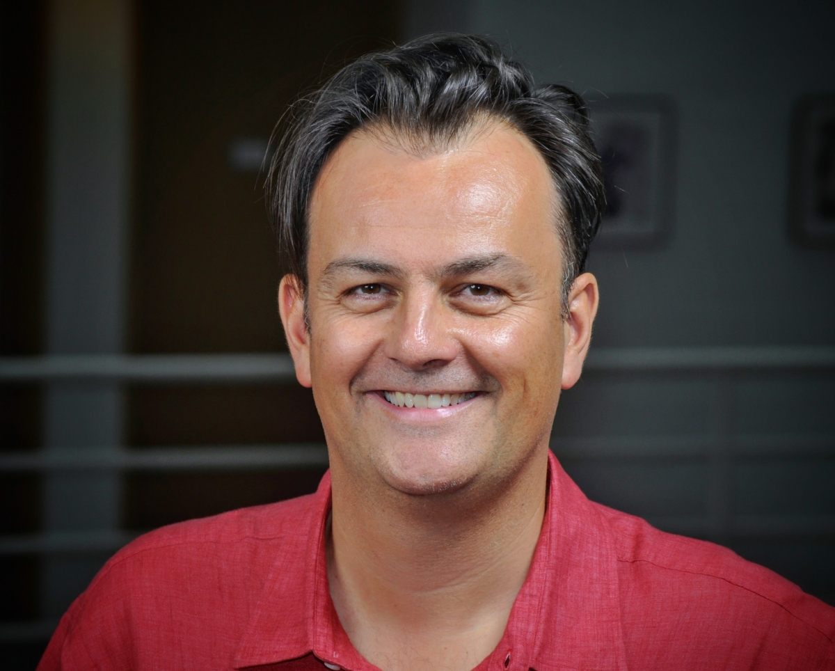 Smiling person with dark hair wearing a red shirt, standing in front of a blurred indoor background.