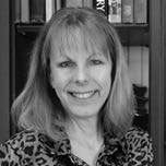 Black and white photo of a person with shoulder-length hair, smiling, wearing a patterned top, standing in front of a bookcase.