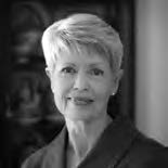 A grayscale portrait of an older woman with short hair, smiling slightly. She is wearing a dark top and looking directly at the camera. The background is softly blurred.