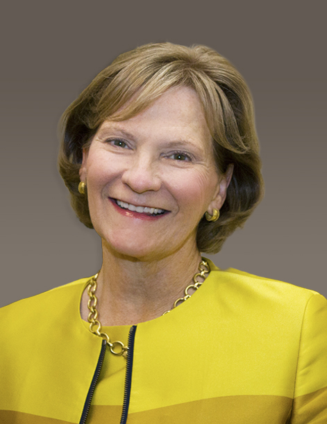 A woman with short, light brown hair smiles warmly. She is wearing a gold necklace and a yellow outfit with gold earrings. The background is a neutral gray-brown gradient.