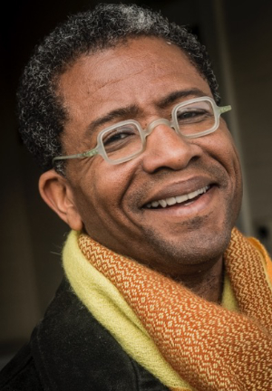 A person with short curly hair and glasses smiles warmly. They are wearing a vibrant yellow and orange scarf and a dark jacket. The background is softly blurred.
