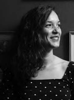 Black and white portrait of a smiling woman with long, curly hair, wearing a polka dot top. She is looking to the side against a dark background with framed pictures partially visible.