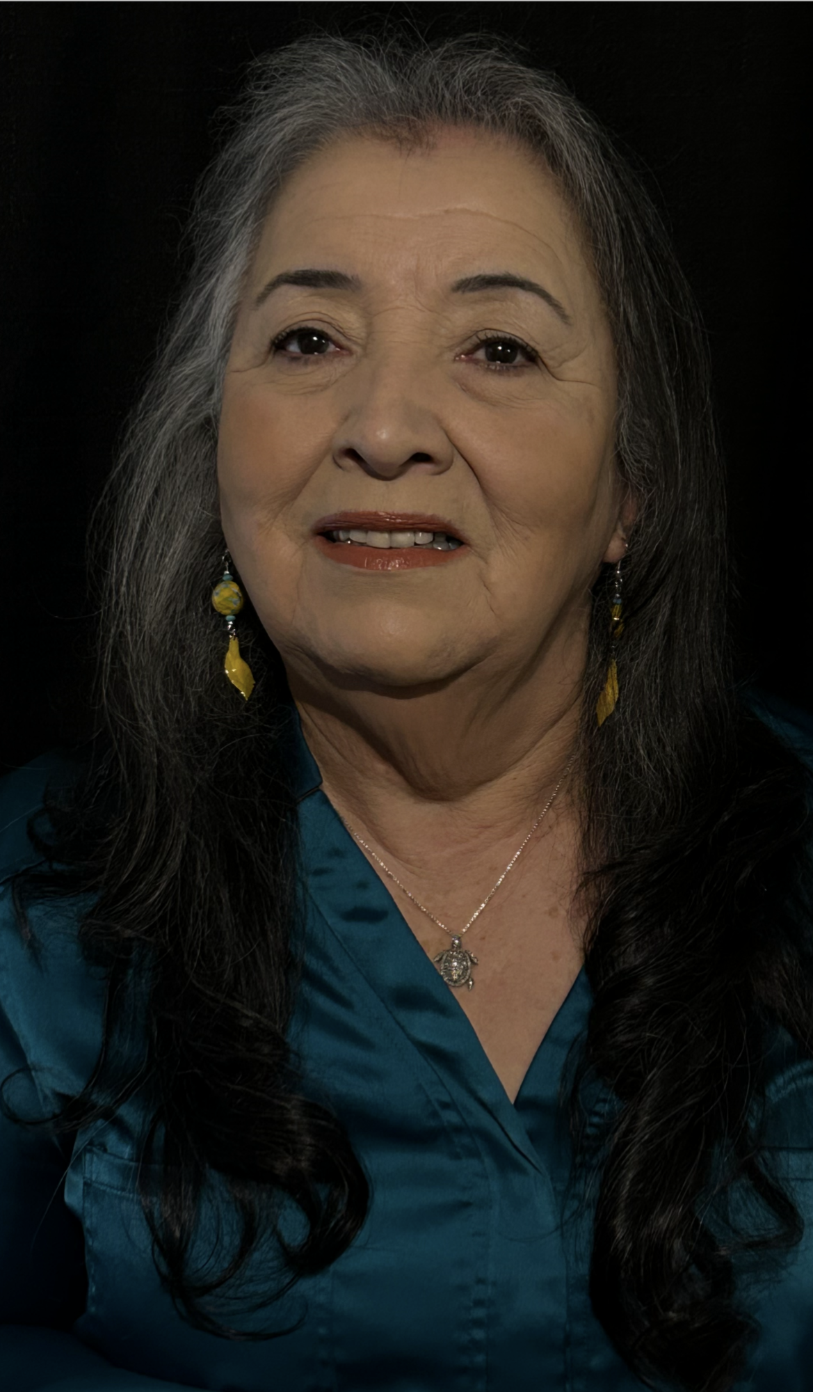 An older woman with long gray hair smiles subtly. Shes wearing a teal blouse and yellow earrings. The background is dark, emphasizing her face.
