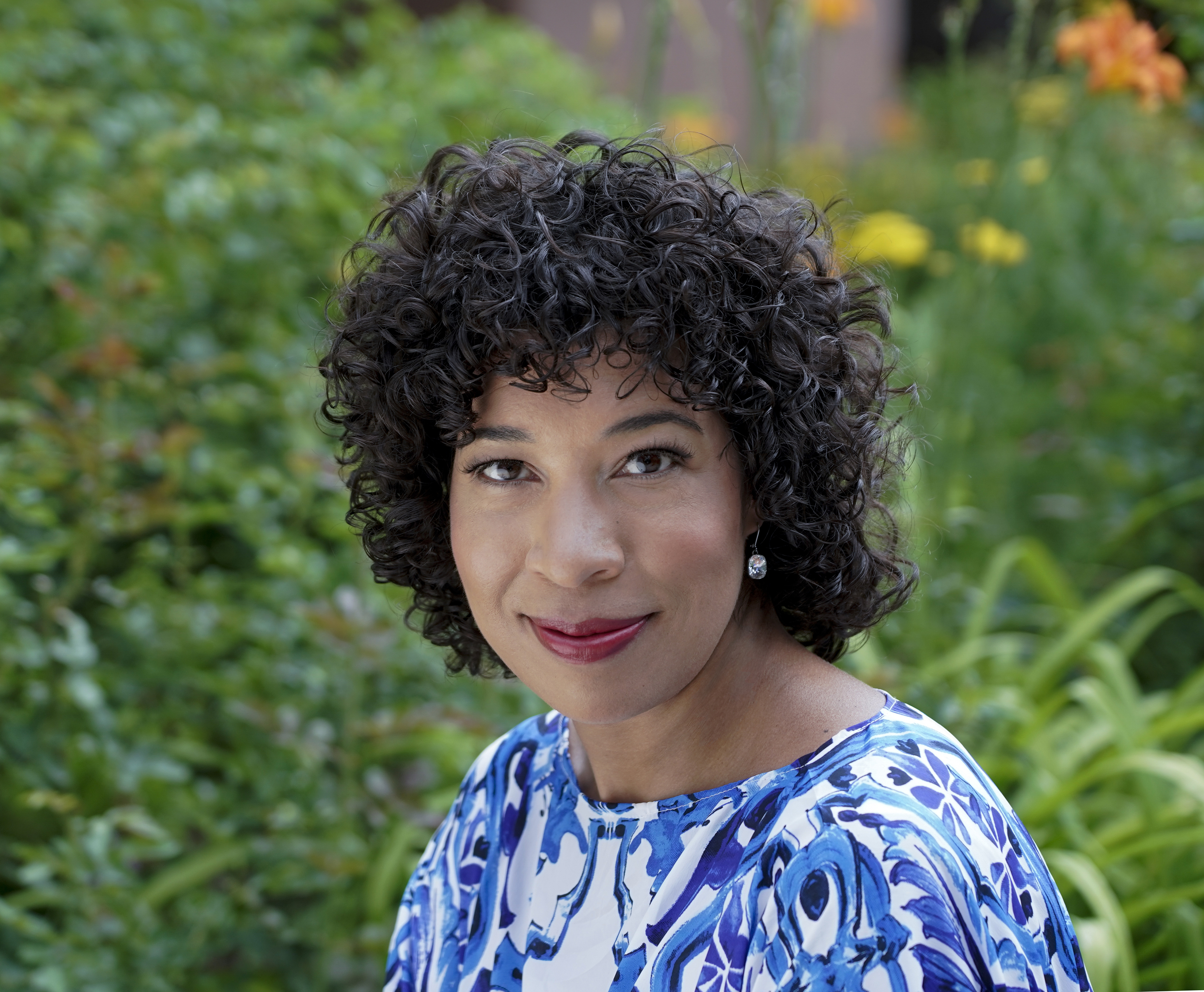 A person with curly hair and a blue patterned top smiles while sitting outdoors. The background is filled with lush greenery and colorful flowers.
