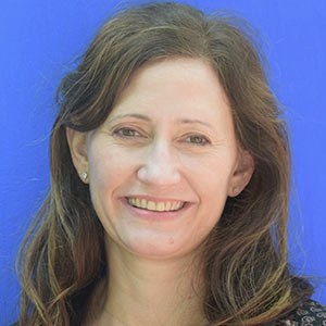 A person with long brown hair smiling in front of a bright blue background. They are wearing a dark top and earrings.