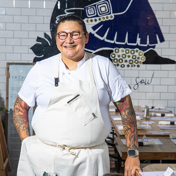 A smiling chef with tattoos stands in a kitchen, wearing a white apron and glasses. White tiles and a black mural are in the background, and tables are set with menus.