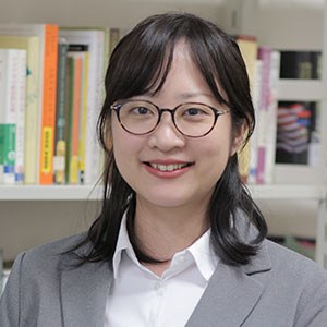 A person with long dark hair and glasses is wearing a light gray blazer over a white shirt, smiling in front of a bookshelf filled with various books.
