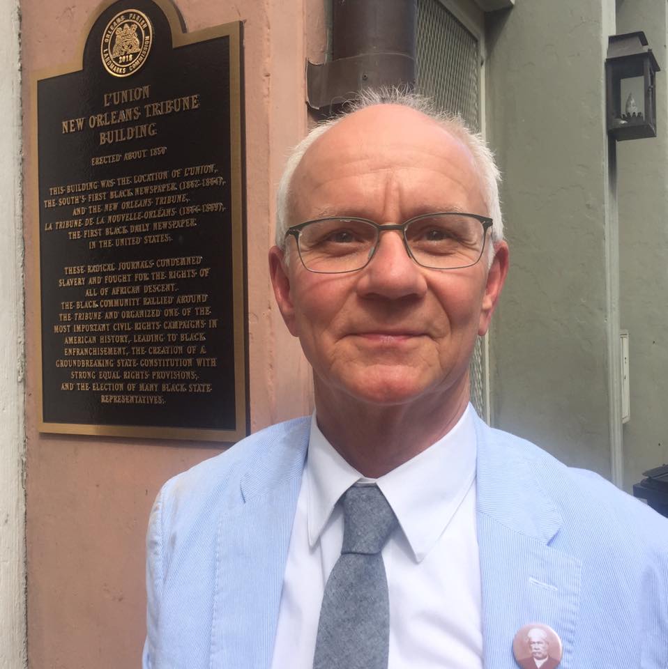 Headshot of Mark Charles Roudane wearing glasses, a light blue suit, and a white shirt standing in front of a historical plaque for the Union New Orleans Tribune Building. The plaque contains text detailing the buildings historical significance.
