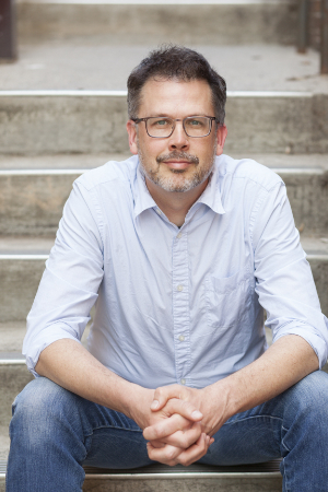 A man with glasses, wearing a light blue button-up shirt and jeans, sits on outdoor steps with hands clasped. He has short, dark hair and a beard. The background features concrete steps.