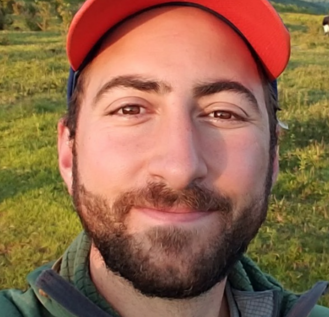 A man wearing a red cap and green jacket smiles while taking a selfie in a grassy, hilly landscape under a clear sky. Trees and shrubs are scattered in the background.