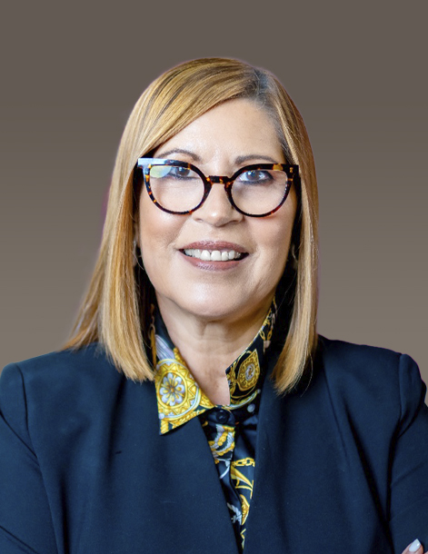 A woman with shoulder-length hair and glasses is smiling. She wears a black blazer over a patterned shirt. The background is a neutral grayish-brown color.