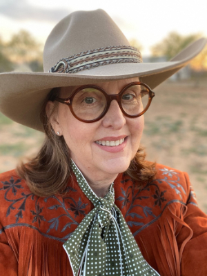 A person wearing a brown cowboy hat, round glasses, and a fringed, embroidered jacket smiles at the camera. The background is a blurred outdoor setting with trees.