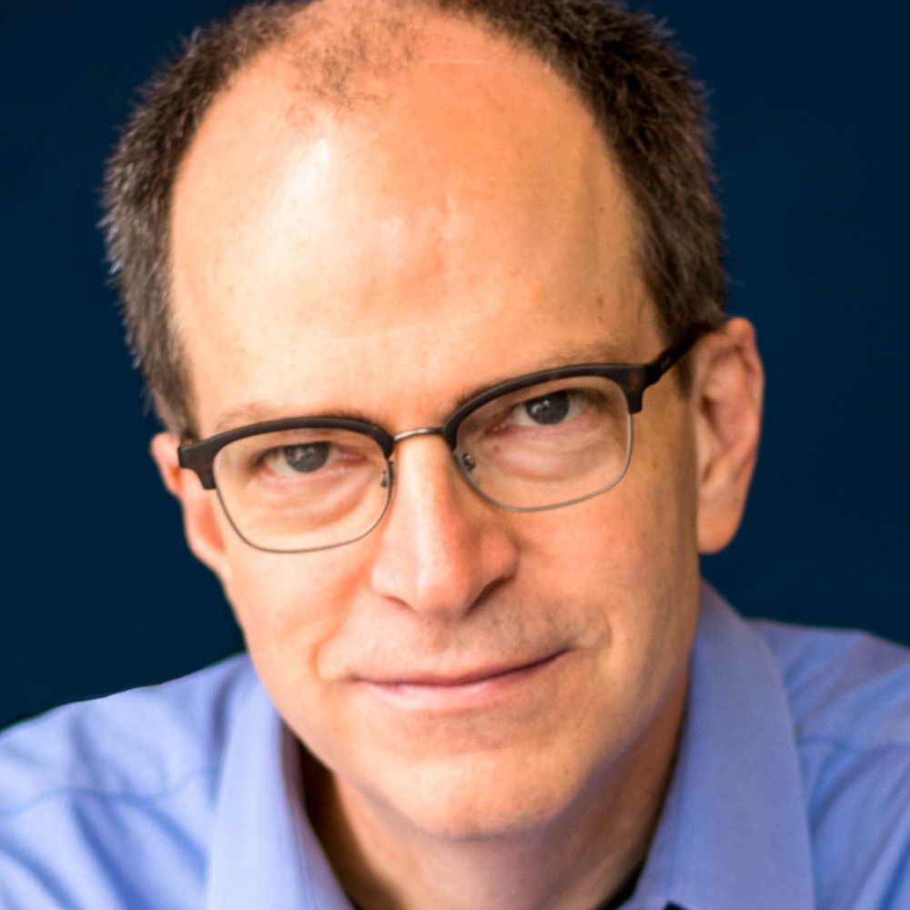 A man with glasses and a receding hairline wears a blue collared shirt, looking directly at the camera against a dark blue background.