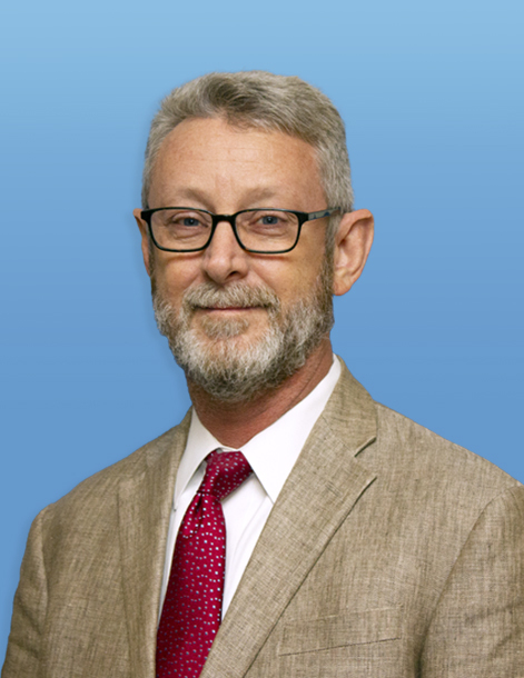 A man with glasses, gray hair, and a beard is wearing a beige suit, white shirt, and red tie. He stands against a plain blue background.