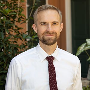 A bearded man in a white shirt and red tie stands outdoors in front of greenery. He has light hair and is smiling, with a building partially visible in the background.