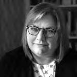 A black and white portrait of a person with shoulder-length hair wearing glasses. They are dressed in a patterned top and a dark jacket, with a background featuring blurred shelves.