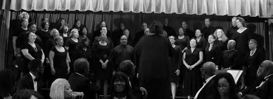 A choir of diverse individuals dressed in black performs on stage with a conductor leading in front. They are positioned against a curtained backdrop, and an audience is partially visible in the foreground.