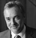 Black and white image of a man wearing a suit and tie. He has short hair and a gentle smile. The background is blurred, focusing attention on his face.