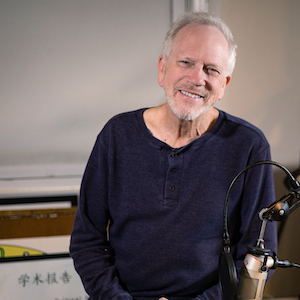 An older man with short white hair and a beard is smiling while seated in front of a microphone. He is wearing a dark long-sleeve shirt. In the background, there are framed pictures leaning against a wall.
