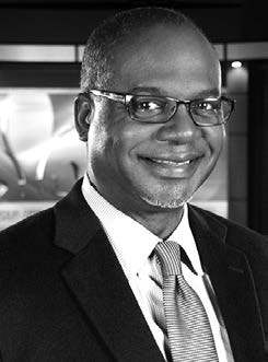 A person is smiling, wearing glasses, a suit, and a tie. The background shows an abstract design. The image is in black and white.