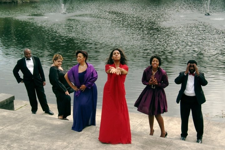 Six people in formal attire pose humorously by a lakeside. One woman in a red dress stands with arms outstretched, while others in purple dresses and black suits strike playful poses. Fountain water sprays in the background.