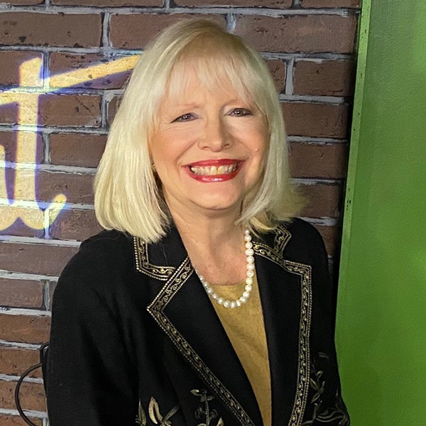 A smiling woman with shoulder-length blonde hair is wearing a black jacket with embroidered details and a pearl necklace. She is standing in front of a brick wall with a hint of neon text visible behind her.