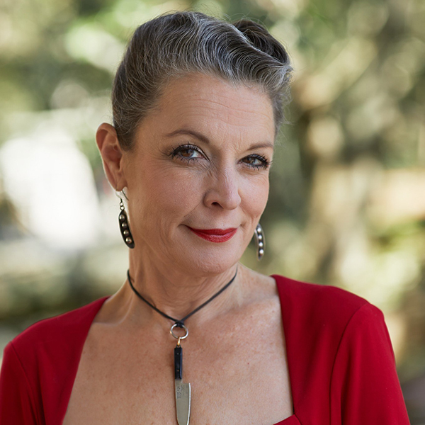 Poppy Tooker with gray hair pulled back, wearing a red top and earrings, smiling slightly. The background is blurred with green foliage, creating a calm and pleasant atmosphere.